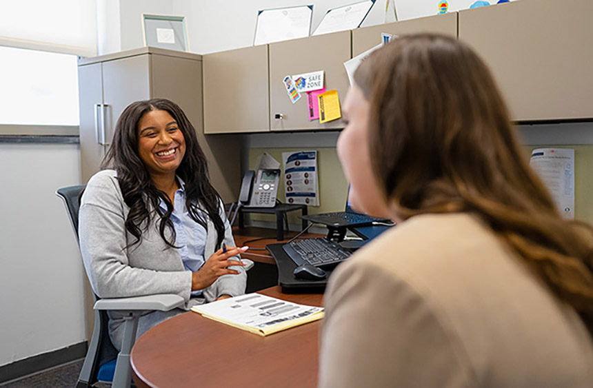 a smiling woman dressed in casual business attire sits at her desk across from a student