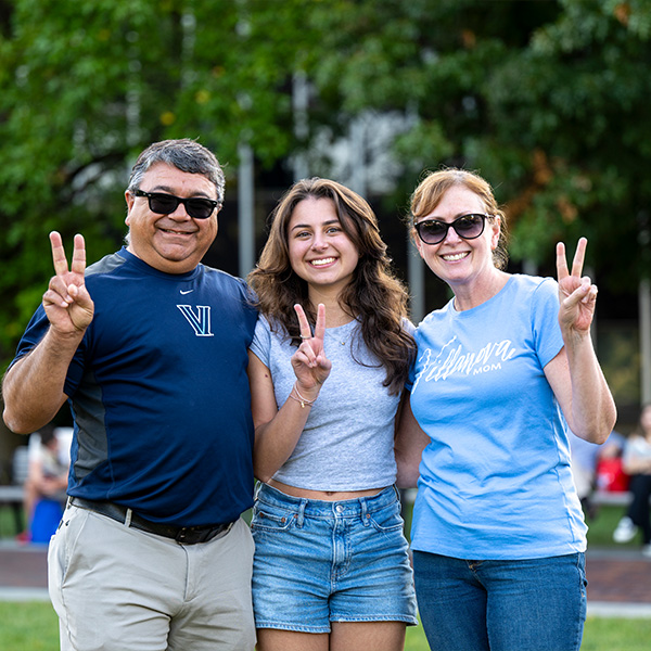 A female student poses with her dad and mom who are all making a V sign with their fingers. 