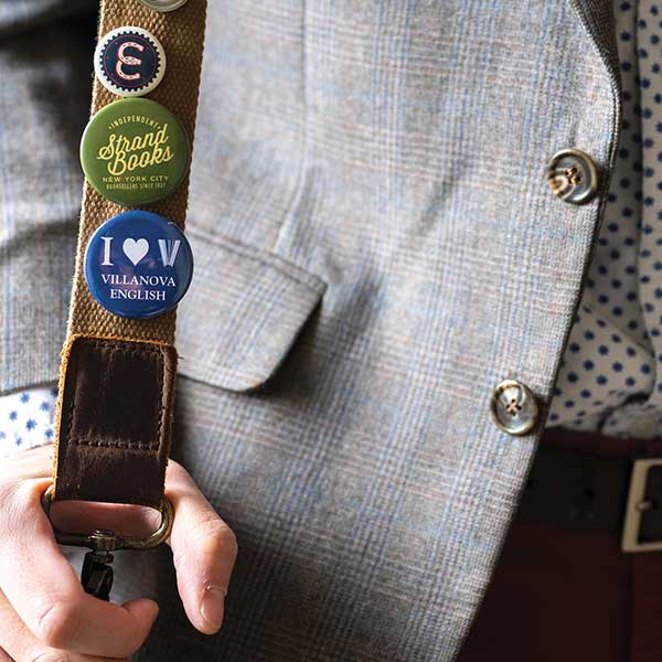 Close-up of a person in a grey suit jacket holding a bag strap with three pins on it.